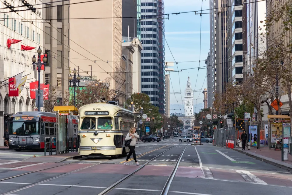 Streetcar and bus stopped at station on Market Street in San Francisco with Ferry Building visible in background.